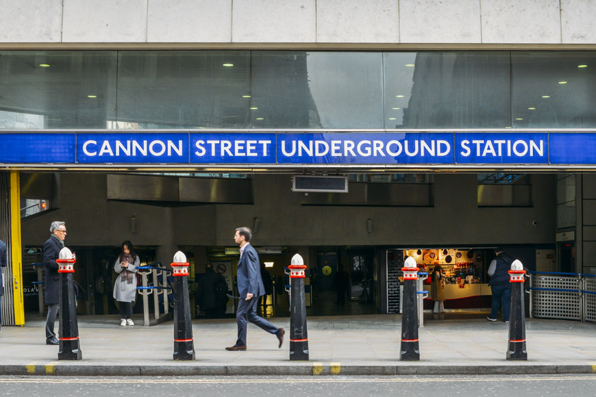 Pedestrians come out of a subway station behind a line of conical black, red, and white bollards