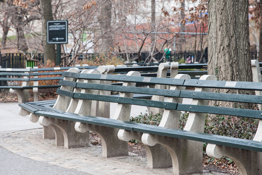 A series of green-slatted wood benches on concrete brackets edge a walkway near a playground