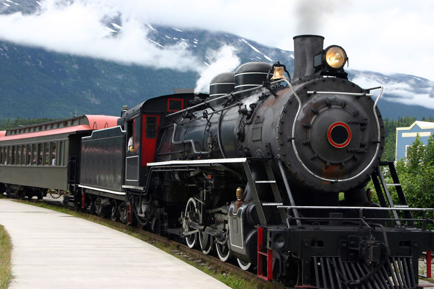 A steam train with steel wheels chugs past a mountain 