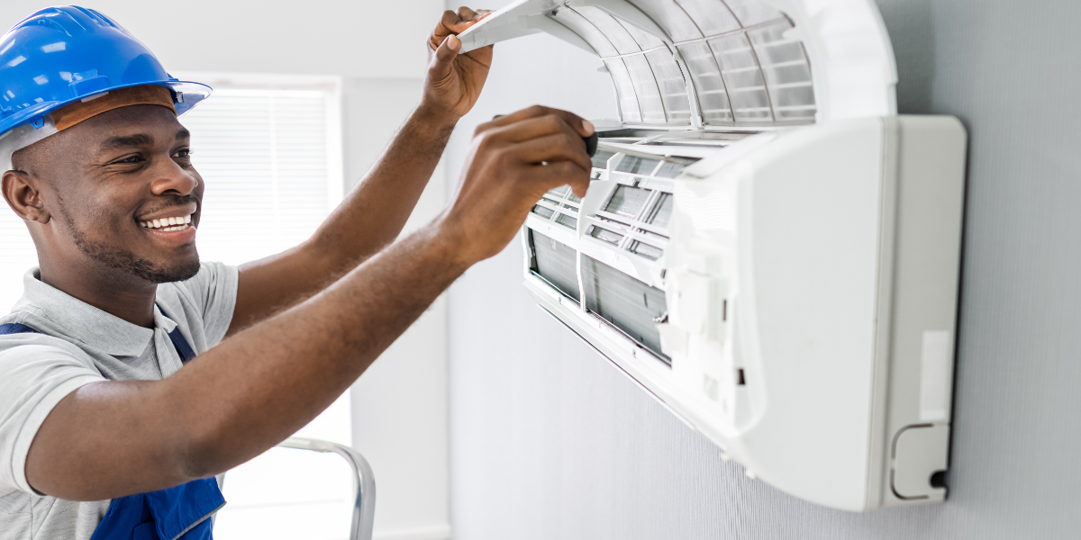 A repair technician, wearing overalls and a hard hat, uses a screwdriver to lift and remove the front panel of an AC unit