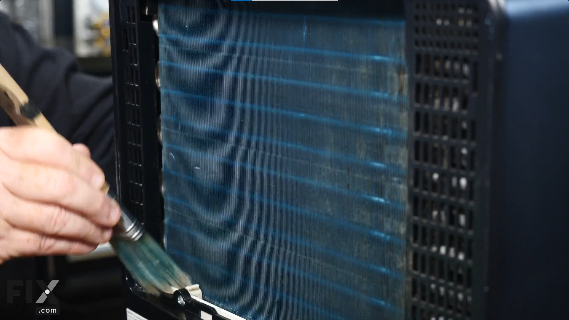 A repair technician sweeping away the dirt particles from the blue coils of a dehumidifier using a soft-bristle brush.