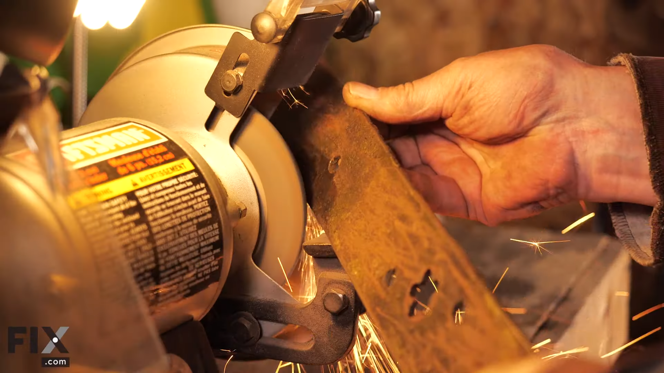 Hand holding rusty lawn mower blade while it sharpens on grinder.