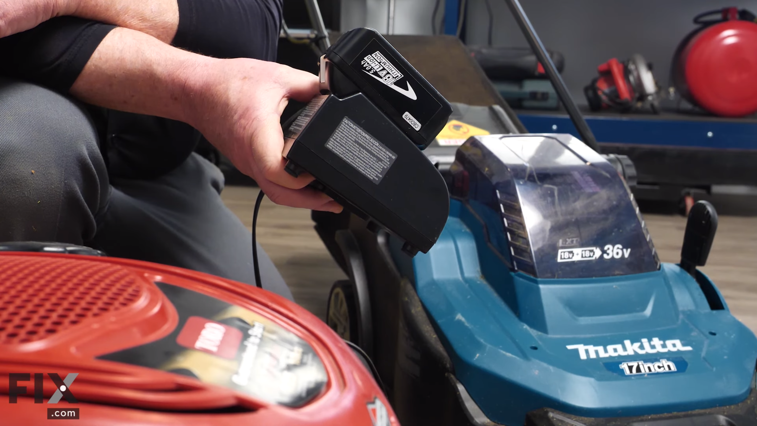 Person holding 18-volt battery in its charger next to two lawn mowers.